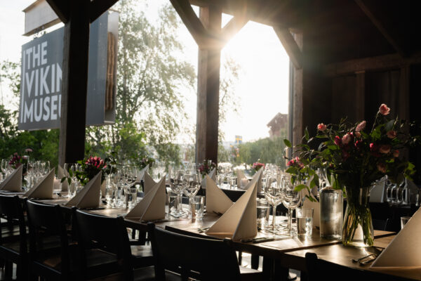 Långbord inne på en restaurang dukade för en tillställning med stora fönster och solsken i bakgrunden.
