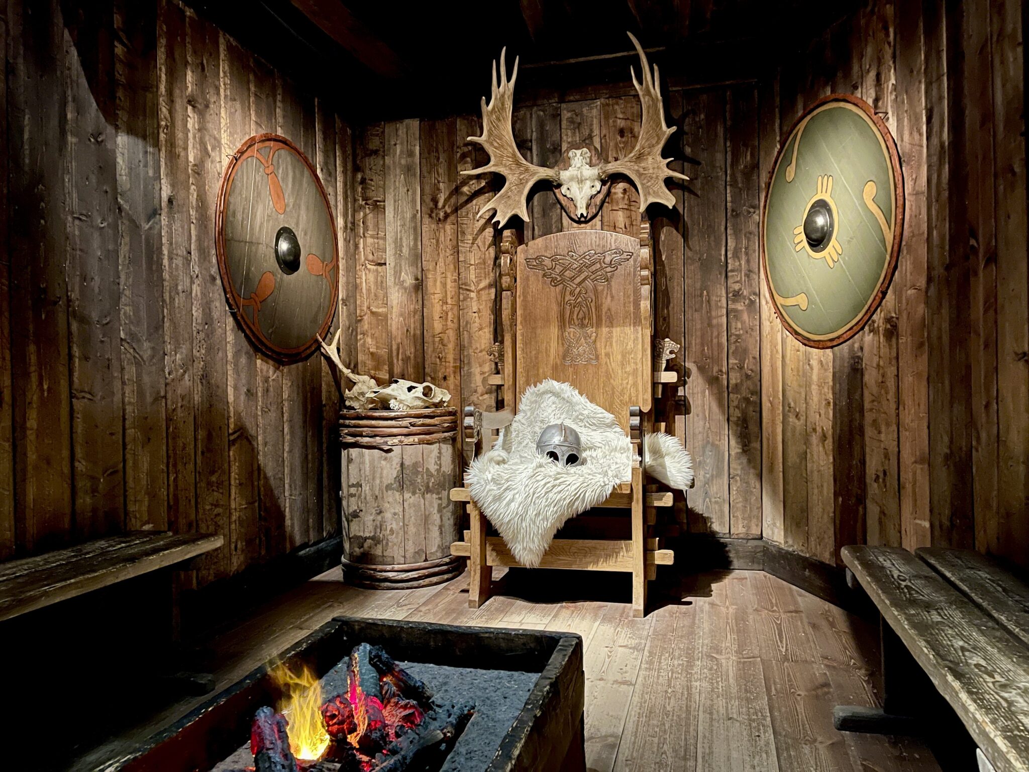 Ett mörkt rum i trä med en hög vikingastol i mitten med en vikingahjälm och en fäll som ligger på och med en brasa framför och sköldar som hänger på väggen.