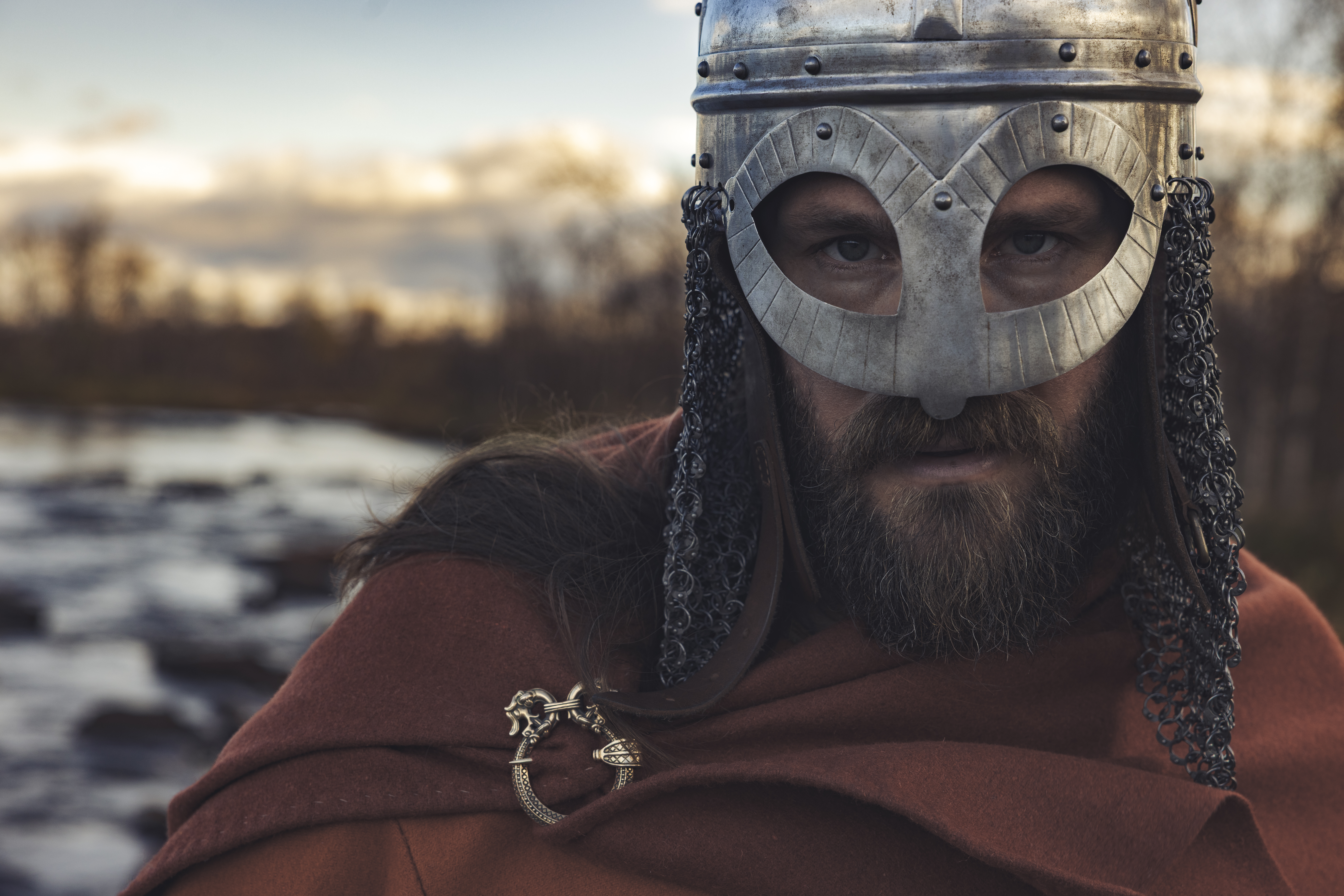 En manlig viking står utomhus med vatten och skog bakom sig iklädd en silverfärgad hjälm.