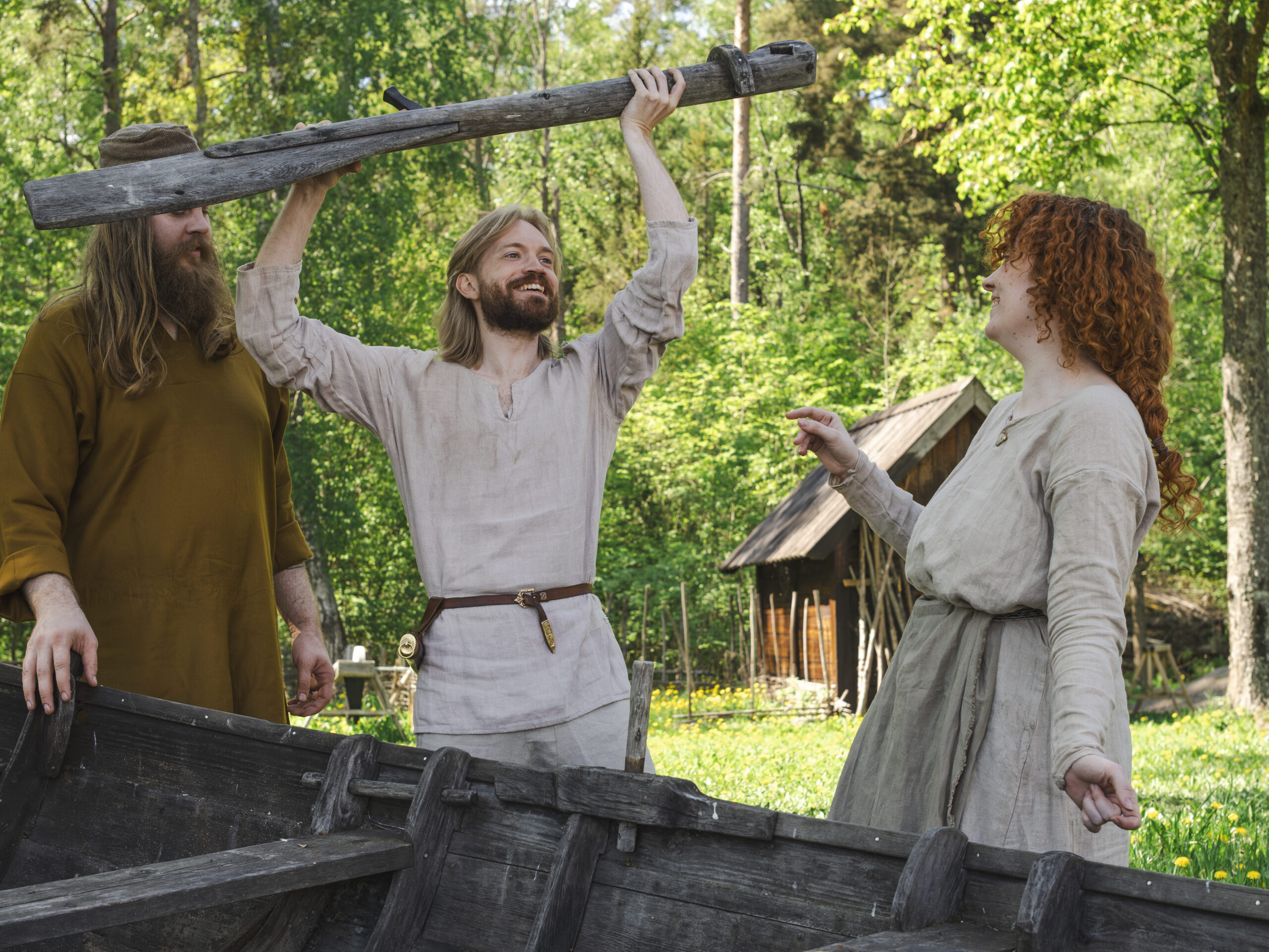 Tre vikingar står ute på gården på sommaren och skrattar tillsammans vid en träbåt.